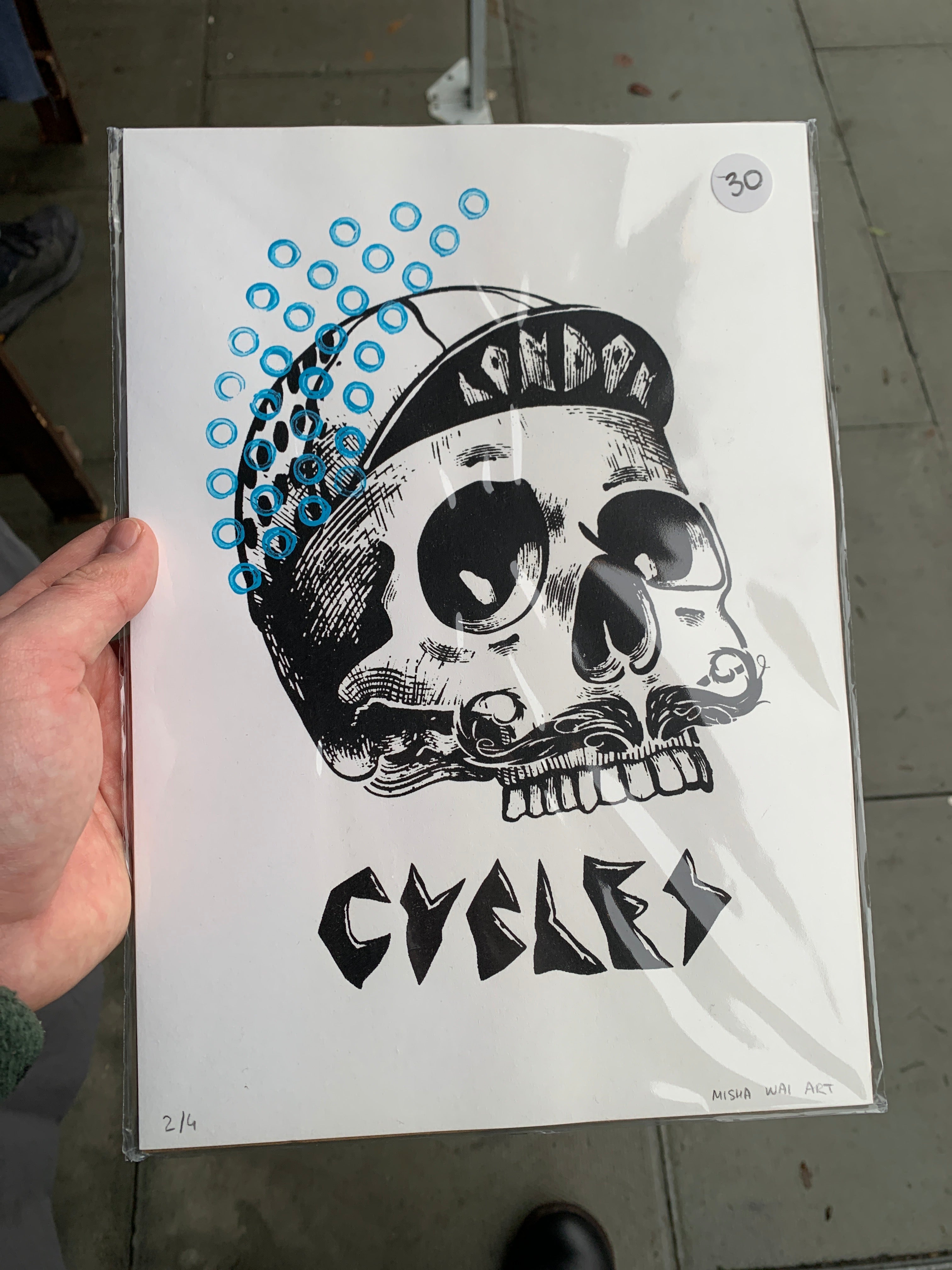 Skull in bike cap and text "Cycles" in black decorated with blue circle patterns on top left in cyan blue printed on white paper.